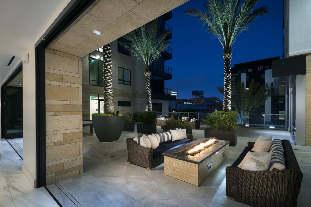 Outdoor patio at night with wicker seating, a rectangular fire pit, and tall palm trees against a dark sky.