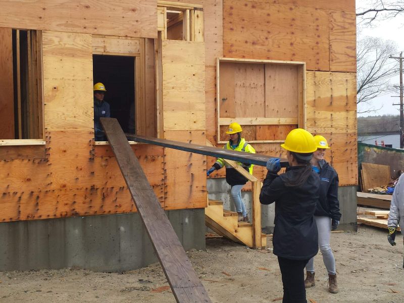 Four people in hard hats move a wooden plank through a window at a construction site.