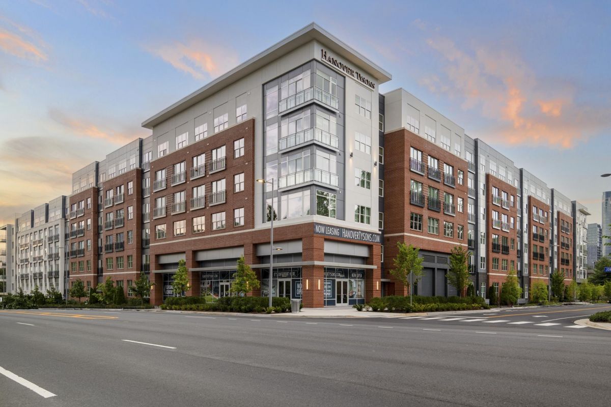 Modern multi-story brick building on a corner, clear sky with light clouds, empty streets and greenery around.