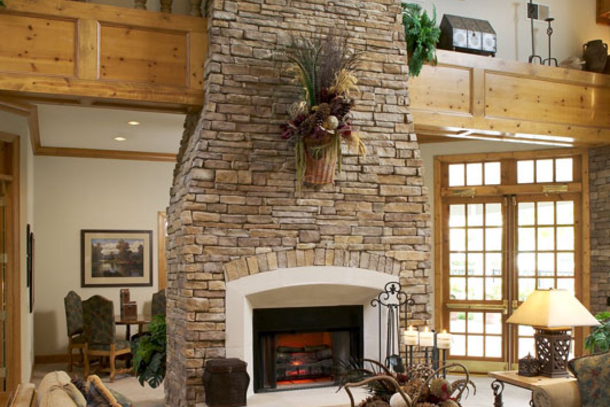 Stone fireplace in rustic living room with wooden beams, beige sofas, and natural light from large windows.