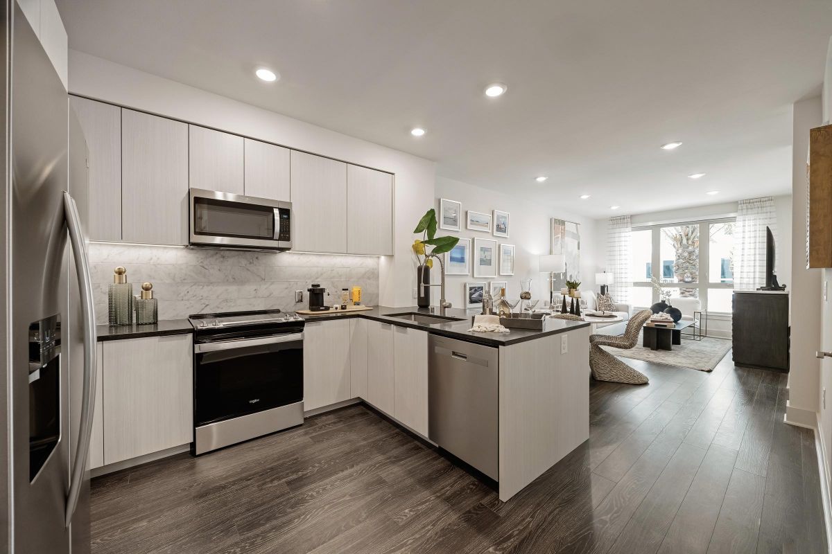 Modern kitchen with stainless steel appliances, marble backsplash, and living room view; wood flooring throughout.