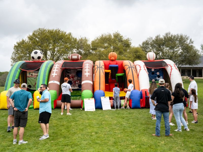 People gather outside near inflatable sports games on a grassy field under a cloudy sky.