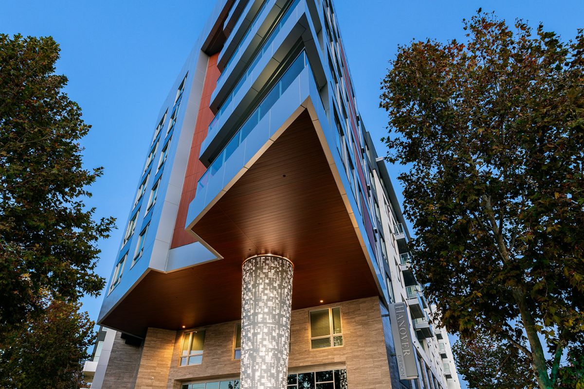 Modern, angular Hanover Diridon apartment building with illuminated pillar at dusk, surrounded by trees and a paved walkway.