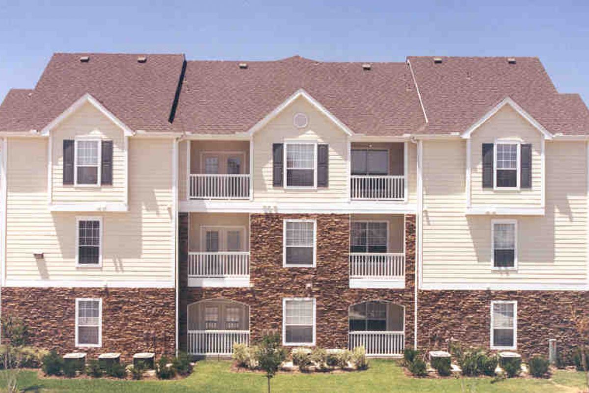 Three-story Dallas apartment building with white siding, stone accents, balconies, and a brown shingled roof.