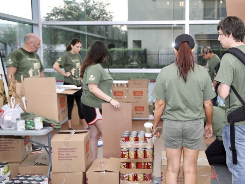 Volunteers in green shirts pack boxes of canned food and supplies inside a building with large windows.