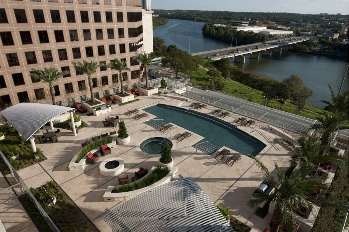 Rooftop pool and lounge area at Ashton Austin with palm trees, fire pit, and river views near city buildings and the highway.
