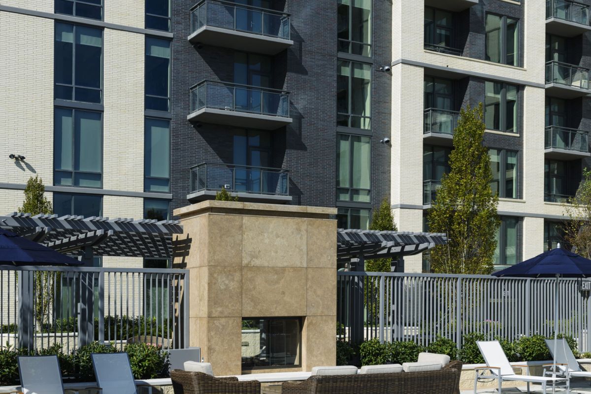 Sunny outdoor pool with lounge chairs, fireplace, and apartment building with balconies in the background.