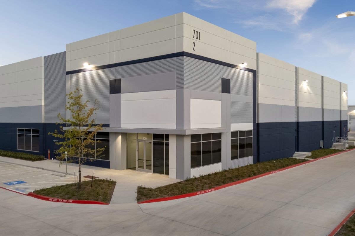 Modern industrial warehouse building with large windows, blue and gray exterior, and an empty parking lot.