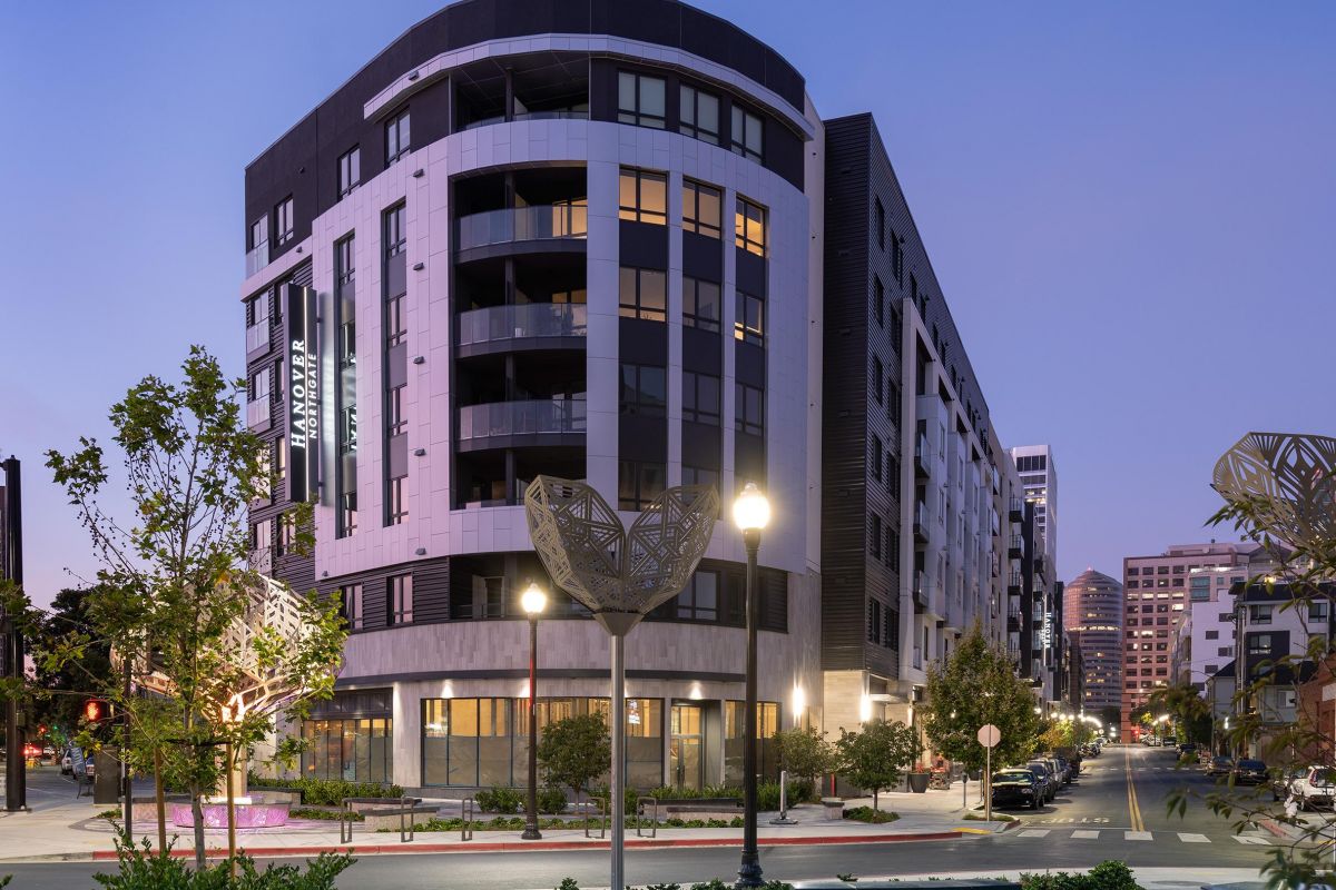 Modern multi-story apartment building on a city corner at dusk, with streetlights and landscaped sidewalks.