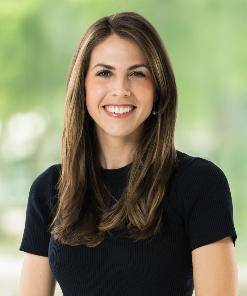 Smiling woman with long brown hair wearing a black top, posed in front of a blurry green background.