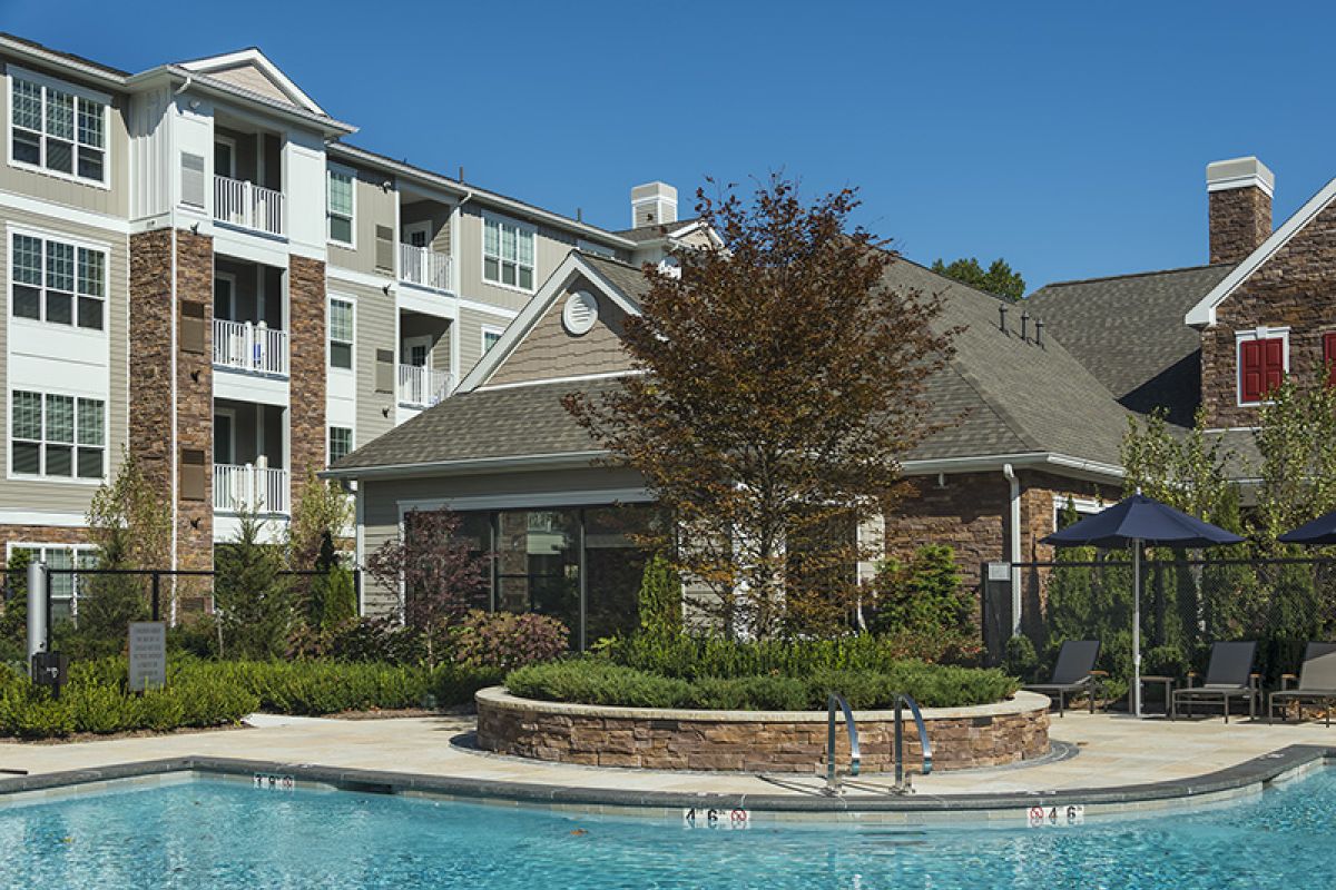 Modern apartment complex with a pool in front, surrounded by landscaping and lounge chairs under umbrellas.