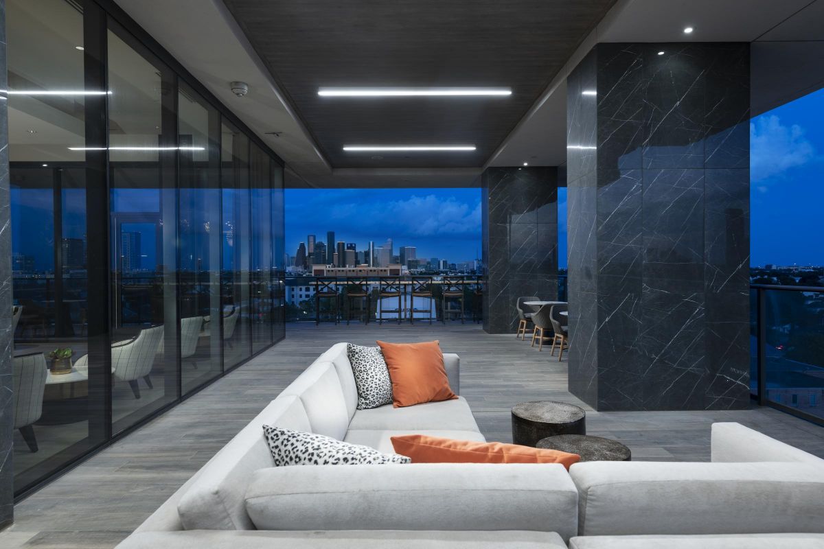 Modern balcony with a sectional sofa, pillows, and city skyline view at dusk through glass walls.