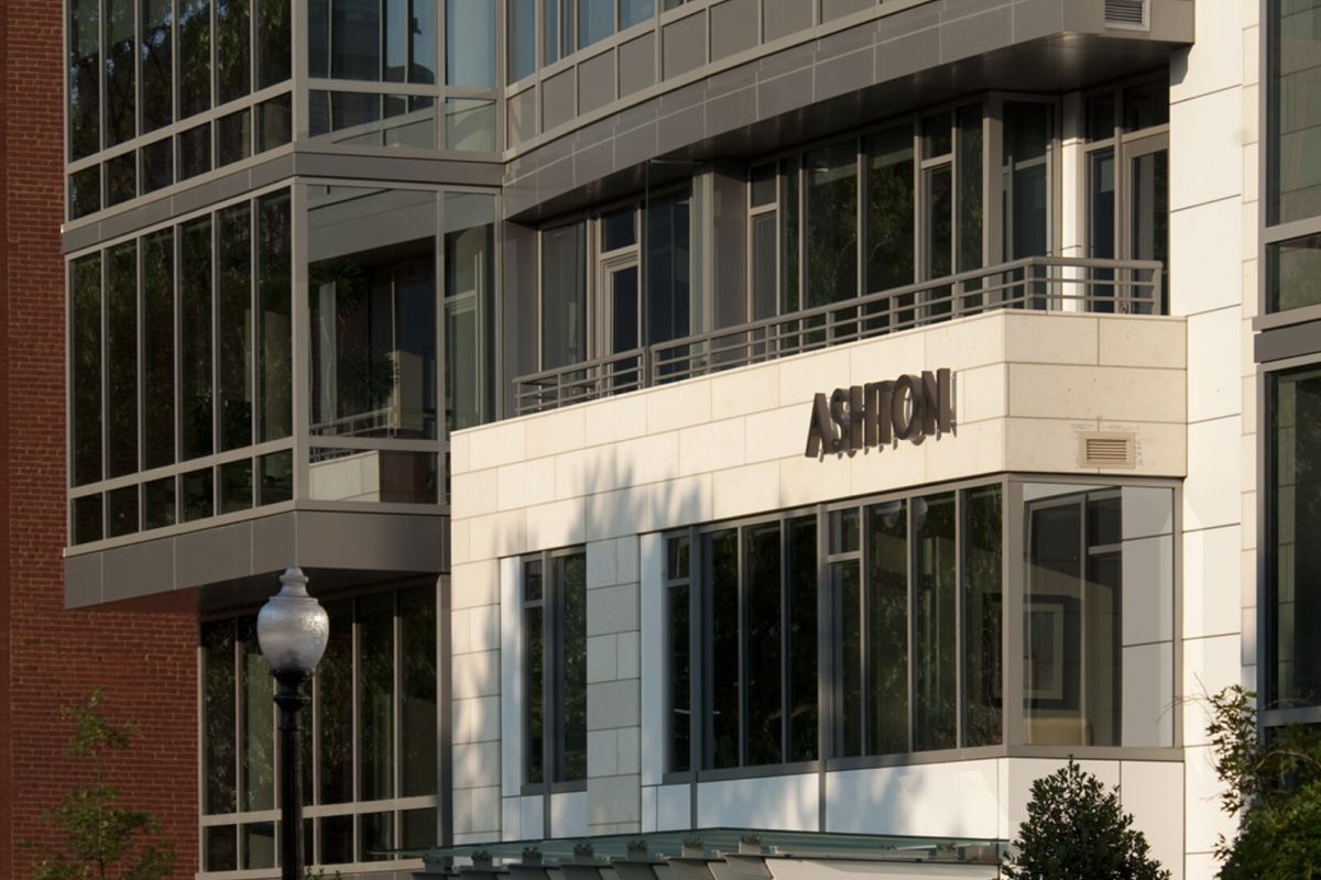 Modern glass and stone apartment building with "Ashton" sign, trees, and a streetlamp in front.