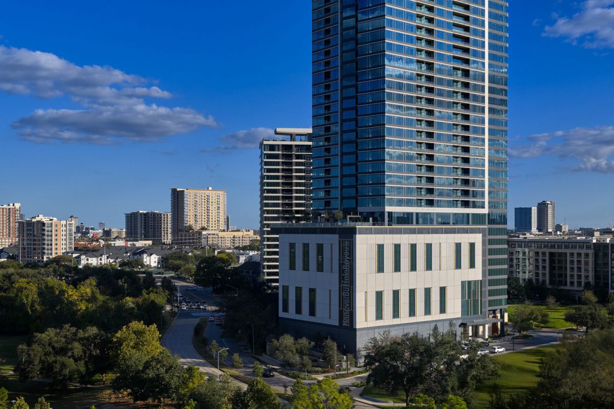 The Hanover skyscraper rises above trees and nearby buildings near Buffalo Bayou under a clear blue sky with scattered clouds.
