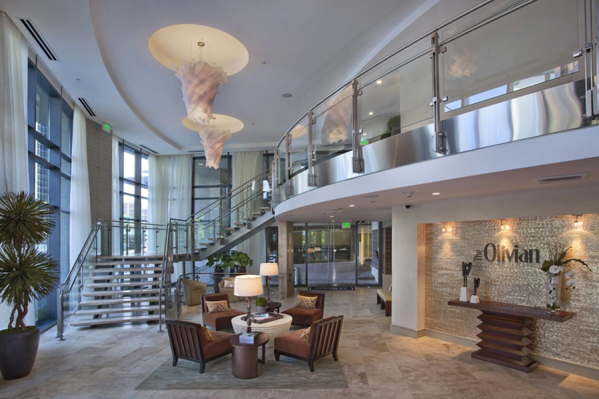 Modern lobby with curved staircase, chandelier, seating area, and “Oman” sign on a textured wall.
