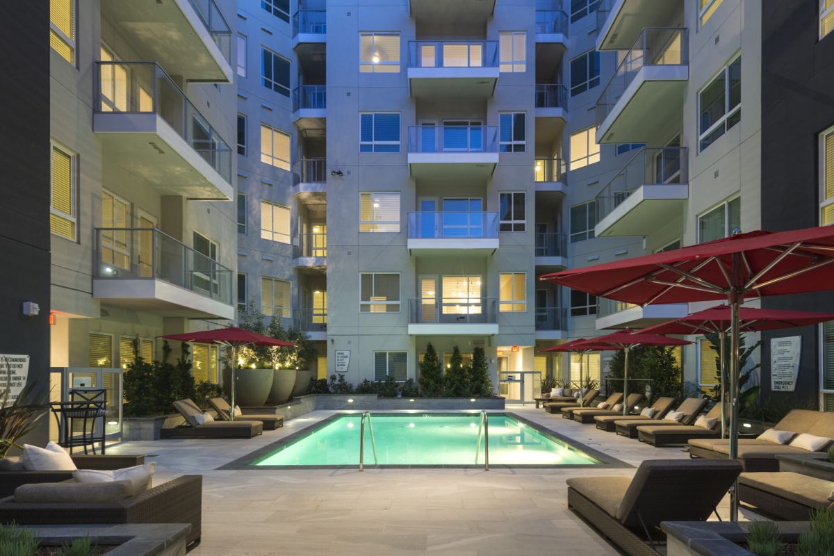 Modern apartment courtyard at dusk with illuminated pool, lounge chairs, and red umbrellas.