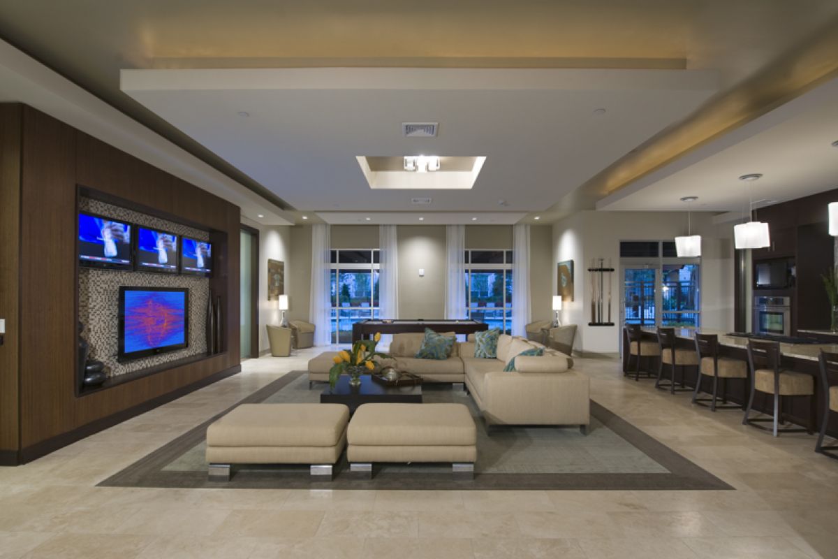 Modern open-concept living room and kitchen in Charles River Landing Newton apartments, featuring beige sofas, bar stools, and wall-mounted TVs.
