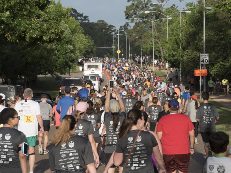 A large crowd of people participates in an outdoor community run or walk event on a tree-lined street.