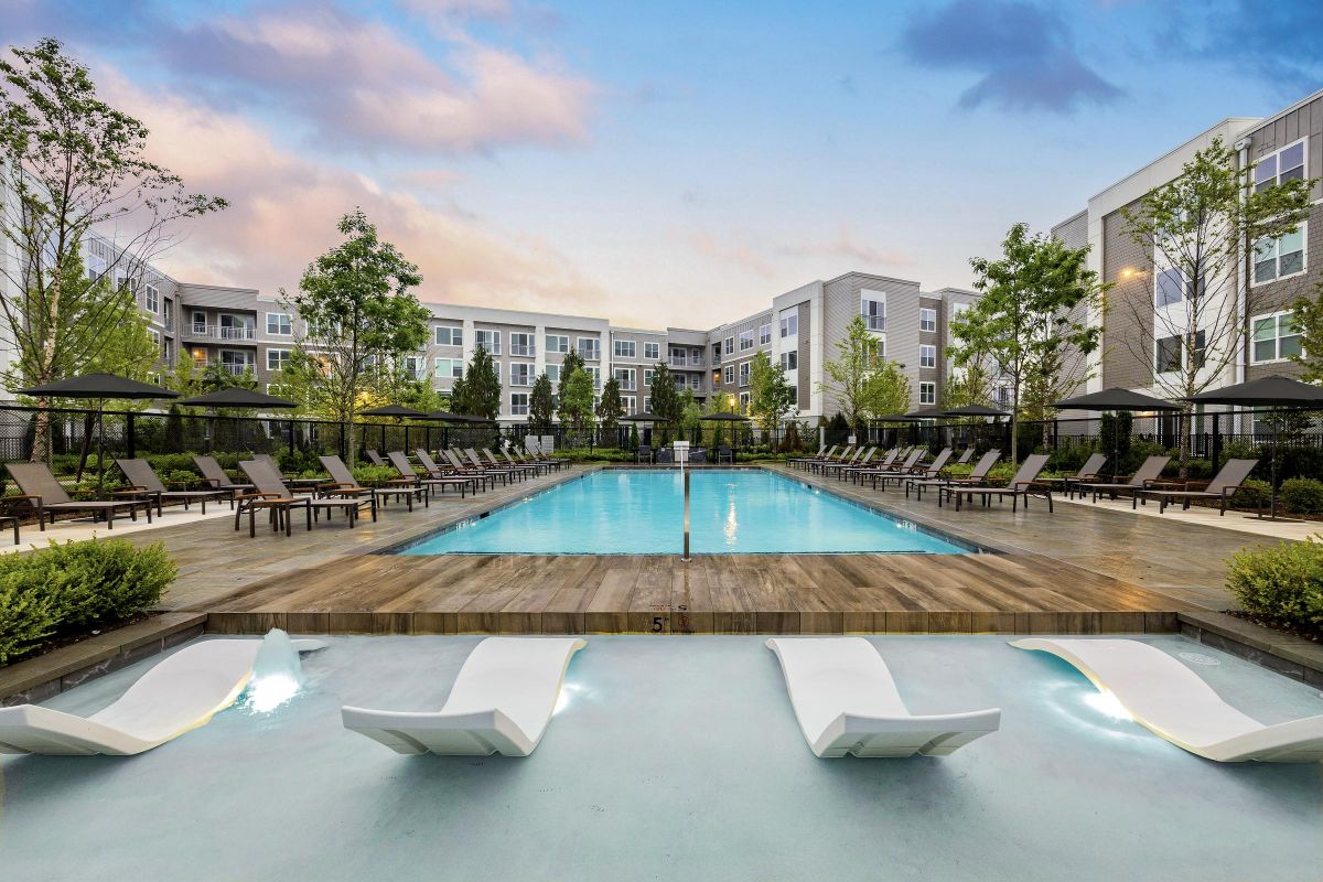 Modern apartment complex with a central swimming pool, surrounded by lounge chairs and trees under a colorful sky.