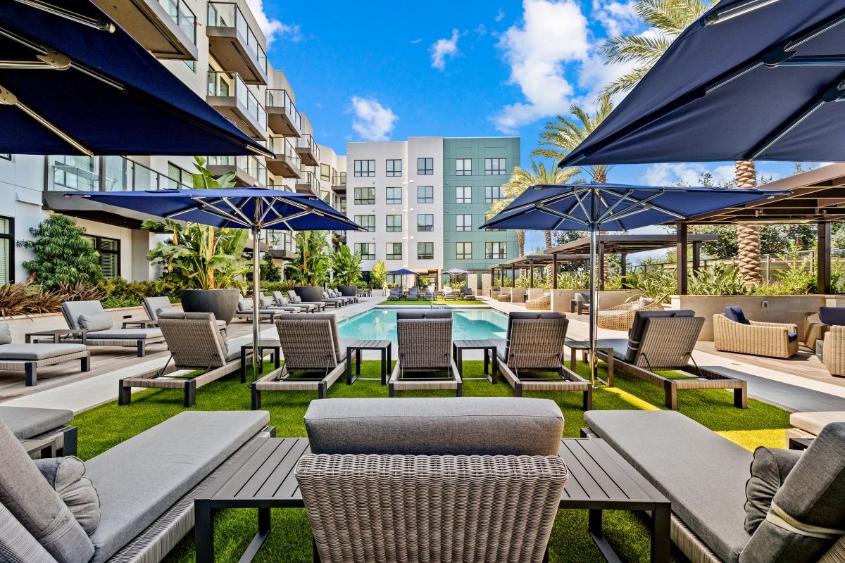 Modern apartment pool area with lounge chairs, umbrellas, and palm trees under a blue sky.