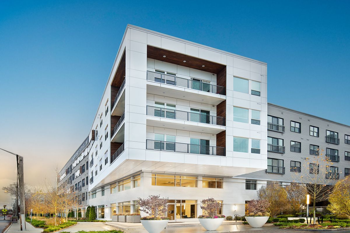 Modern white Hanover Mystic River apartment building with balconies, large windows, and landscaped entrance under a blue sky.