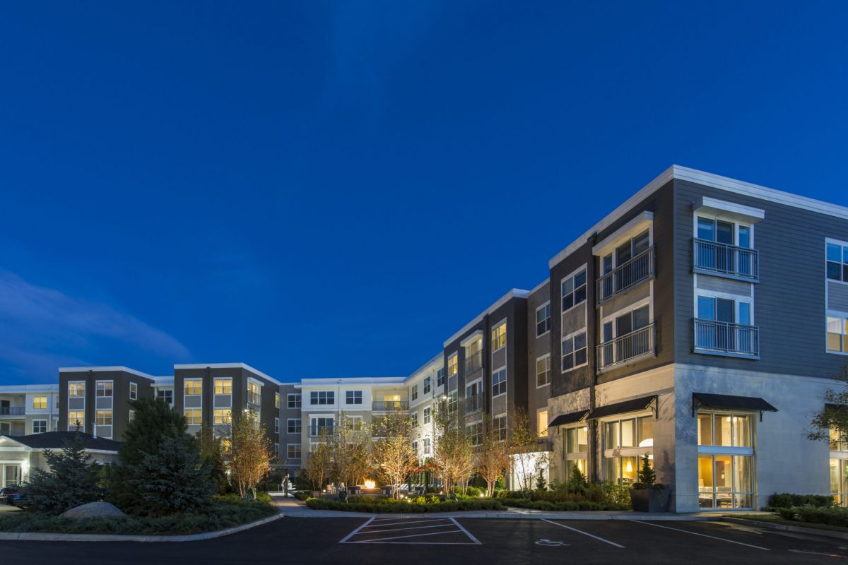 Modern apartment buildings with lit windows and landscaped grounds at dusk under a deep blue sky.