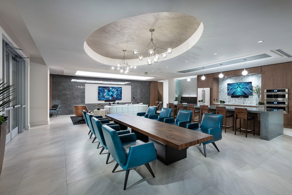 Modern lounge at Hanover Diridon with a long wooden table, blue chairs, a kitchen area, and stylish lighting fixtures.