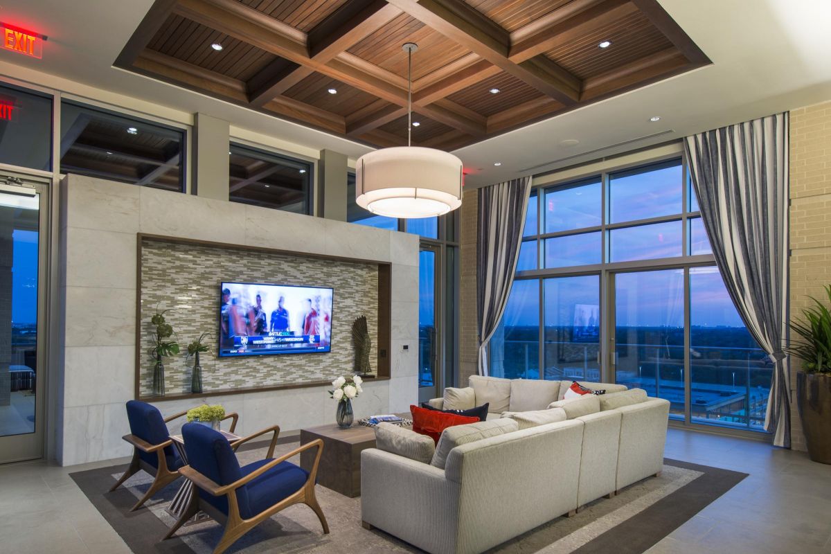 Hanover Company Modern living room with a large TV, gray sectional sofa, blue chairs, and a wood-paneled ceiling. Floor-to-ceiling windows.