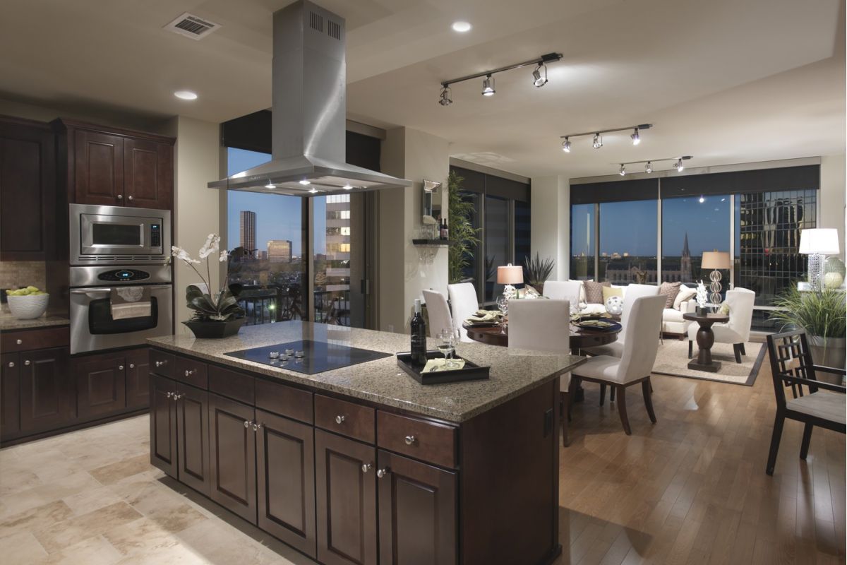 Modern kitchen with island, open to dining and living area, large windows showing a city view at dusk.