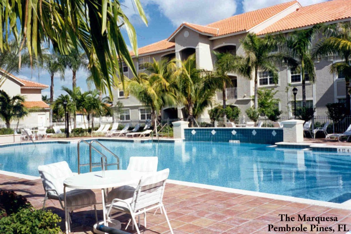 Sunny outdoor pool with white chairs, palm trees, and Broadwater Galveston hotel in the background.