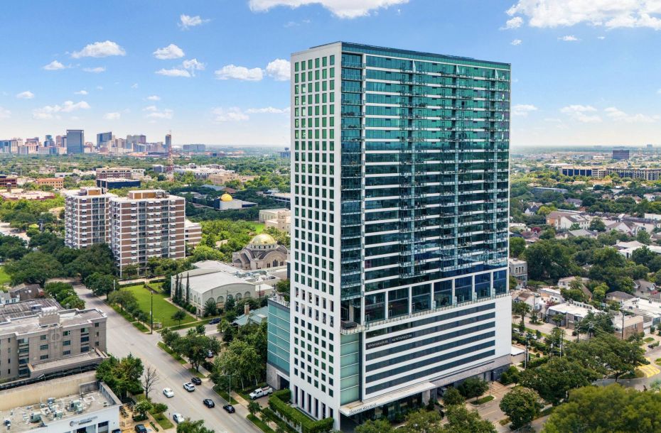 Hanover Company, Hanover Montrose Tall building in a cityscape with green-tinted windows, surrounded by trees and other buildings under a blue sky.