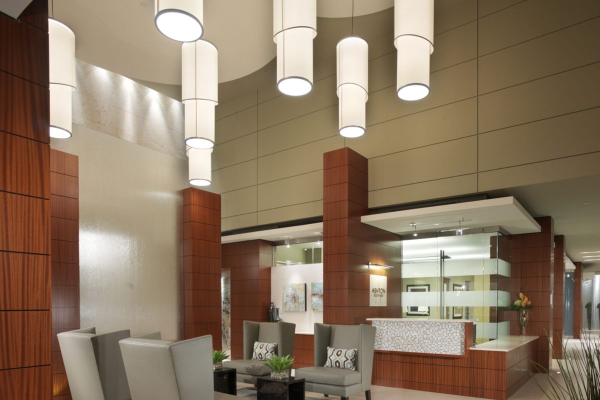 Modern hotel lobby with tall cylindrical lights, gray chairs, brown columns, and a reception desk in the background.