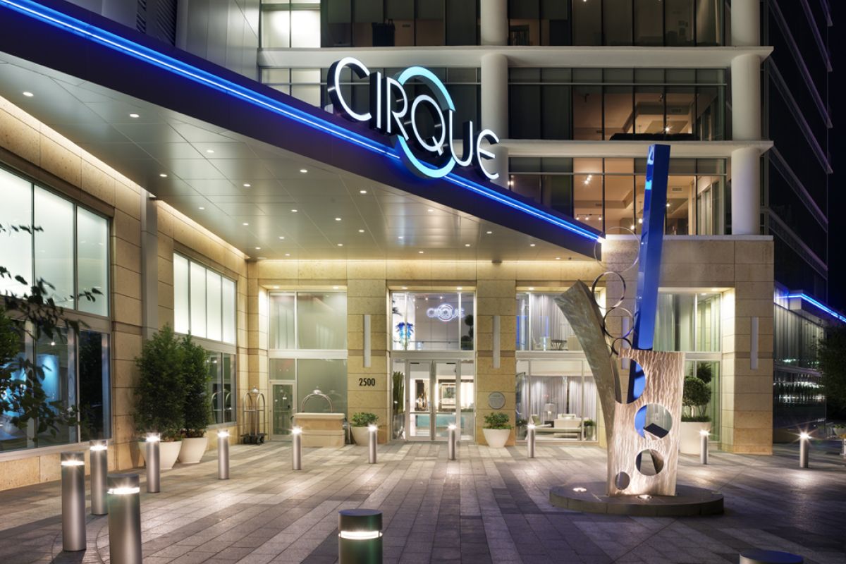 Modern building entrance at night with "CIRQUE" sign, blue lighting, and metal sculpture in front.