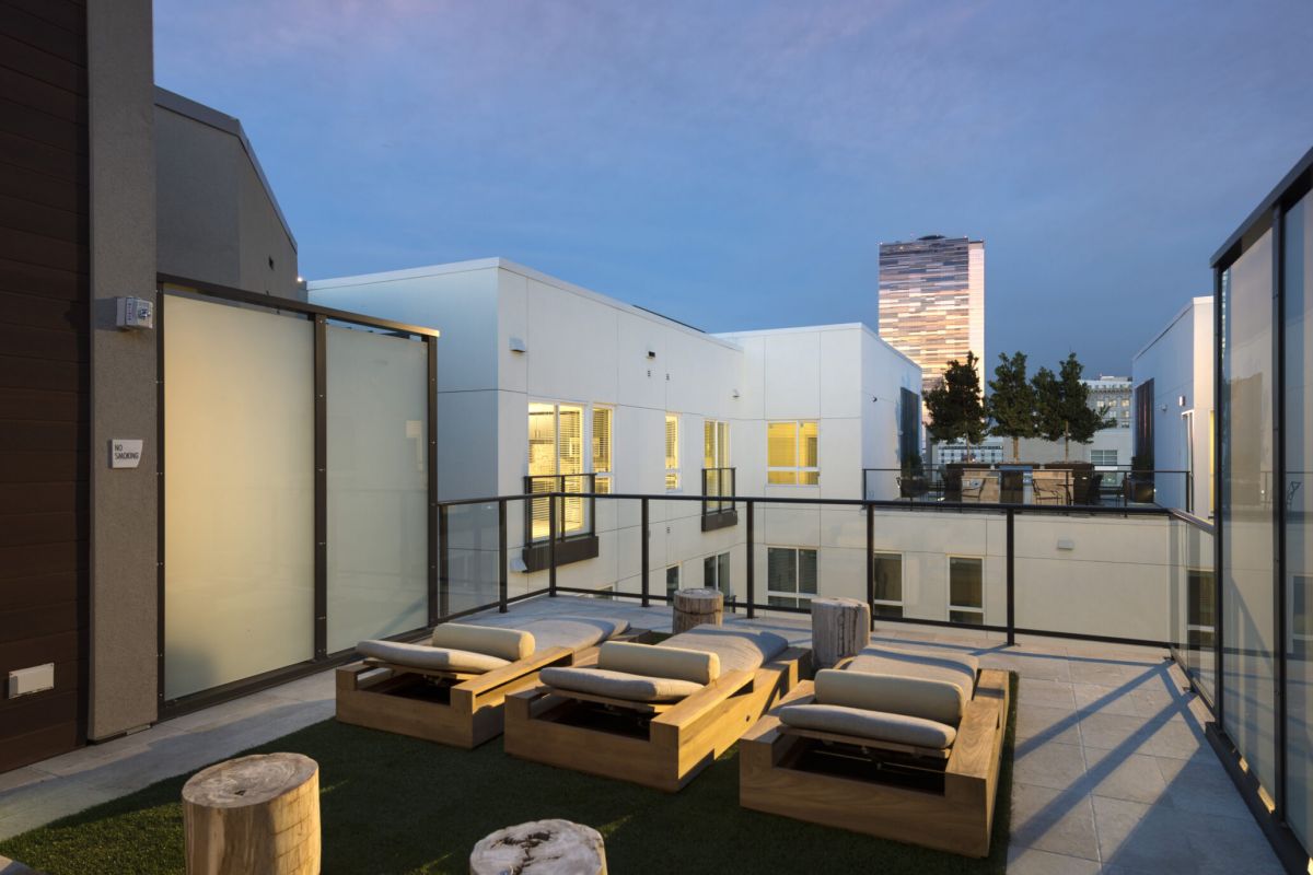 Modern rooftop lounge with wooden seating, glass railings, and city buildings at sunset.