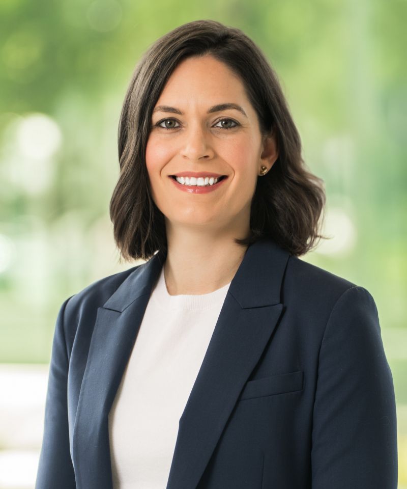 Steffi Russell, a woman with dark hair in a navy blazer and white top, smiles against a blurred green background.