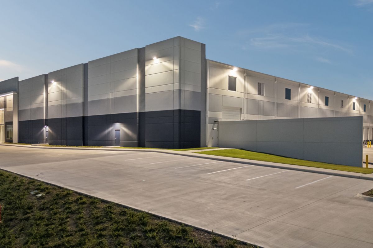 Modern industrial warehouse building with exterior lights and an empty parking lot at dusk.