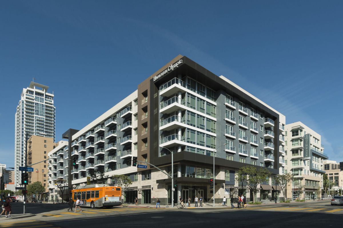 Modern mid-rise apartment building on a city corner with people walking and a bus driving by on a sunny day.