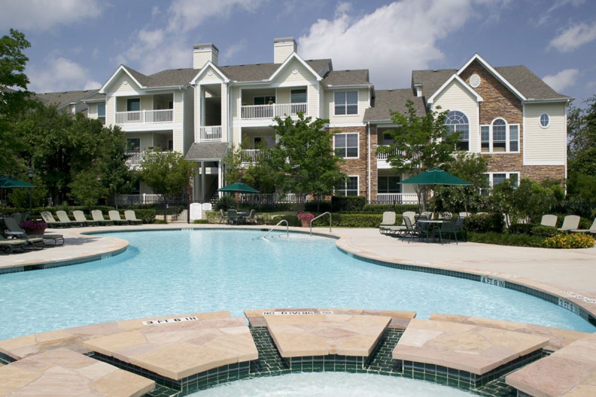 Apartment complex with balconies and a swimming pool surrounded by lounge chairs and umbrellas on a sunny day.