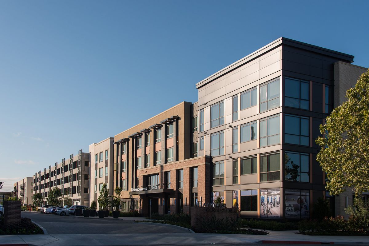 Modern apartment complex with four stories, large windows, and cars parked along the street on a clear day.