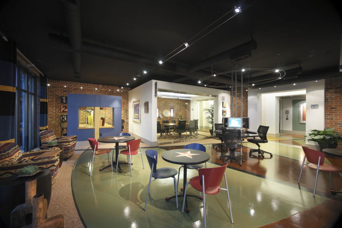 Modern office lounge with colorful chairs, round tables, and exposed brick walls under ambient lighting.
