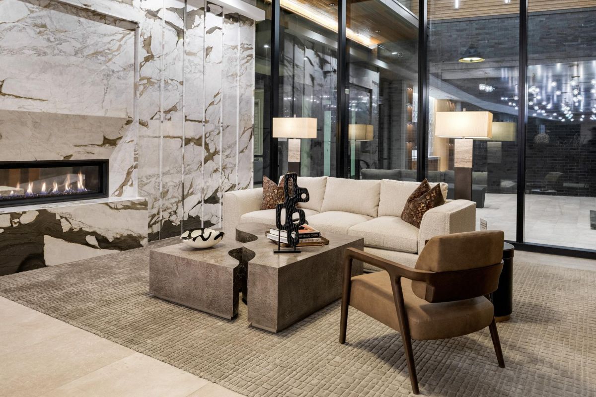 Modern living room with marble fireplace, beige sofa, armchair, and large glass windows overlooking a patio.