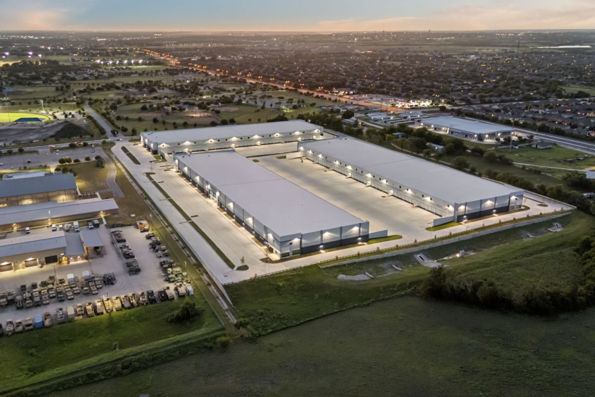 Aerial view of a large industrial complex with multiple warehouses lit up at dusk, surrounded by greenery.
