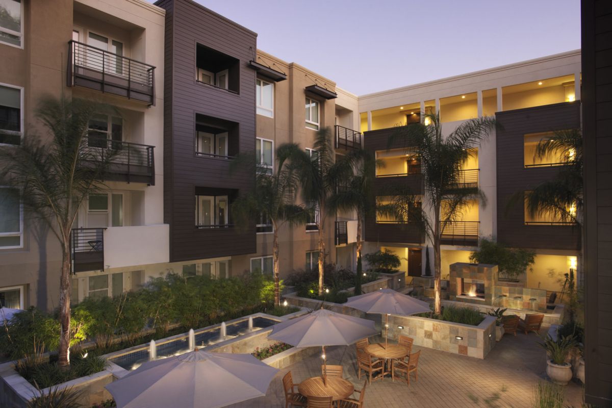 Modern apartment courtyard at dusk with palm trees, outdoor seating, umbrellas, and warm lighting.