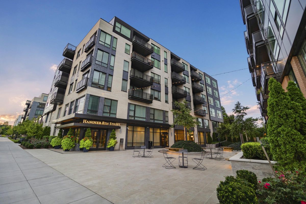 Modern apartment building with balconies, surrounded by trees and outdoor seating under a clear blue sky.