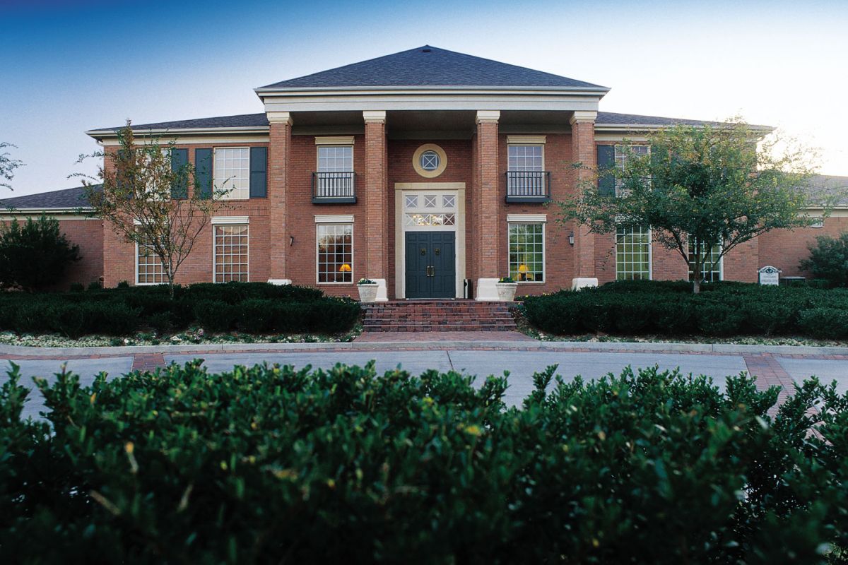 A large brick building with columns, tall windows, and a double-door entrance, surrounded by bushes and trees.