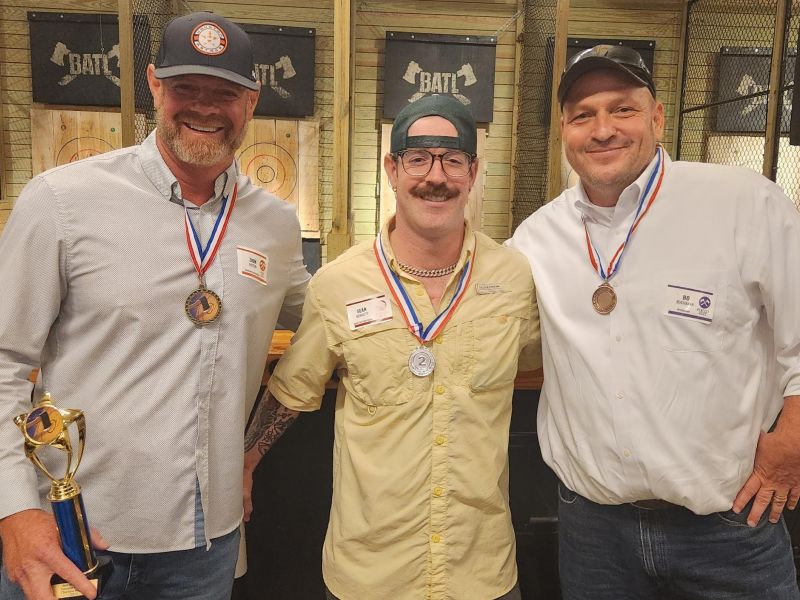 Three men wearing medals pose and smile indoors; one man is holding a trophy.