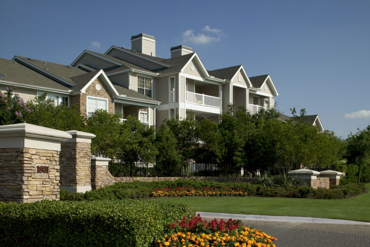 Modern apartment building with balconies, surrounded by lush landscaping and colorful flowers under a clear sky.