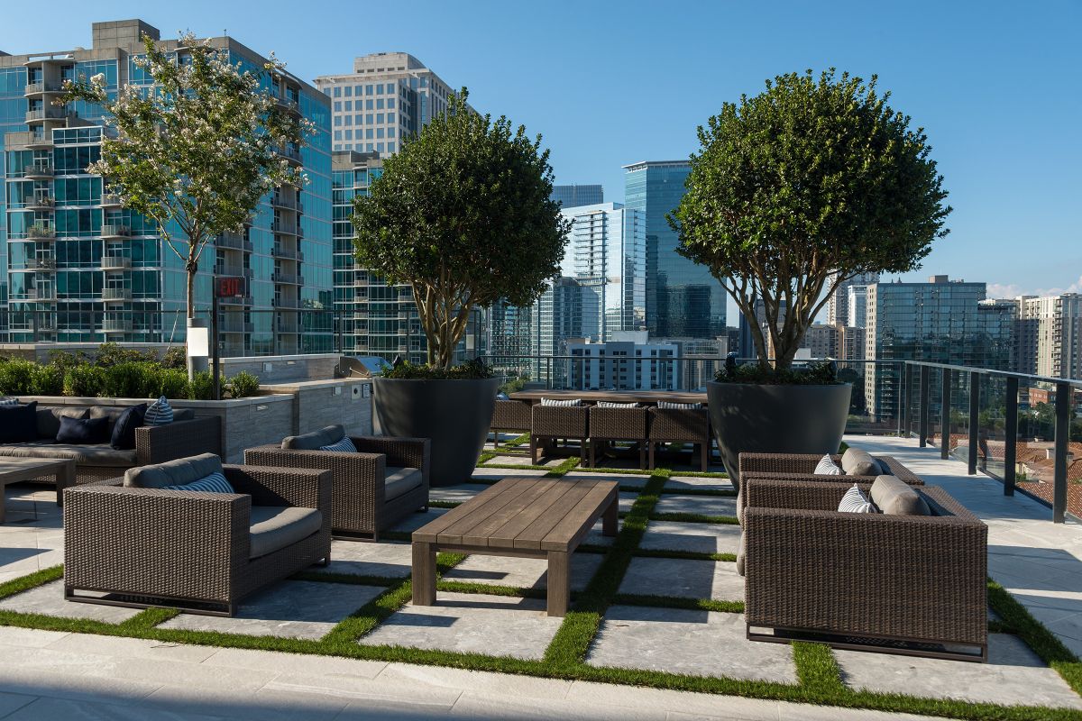 Rooftop patio with wicker sofas, wooden tables, potted trees, and city skyscrapers in the background.