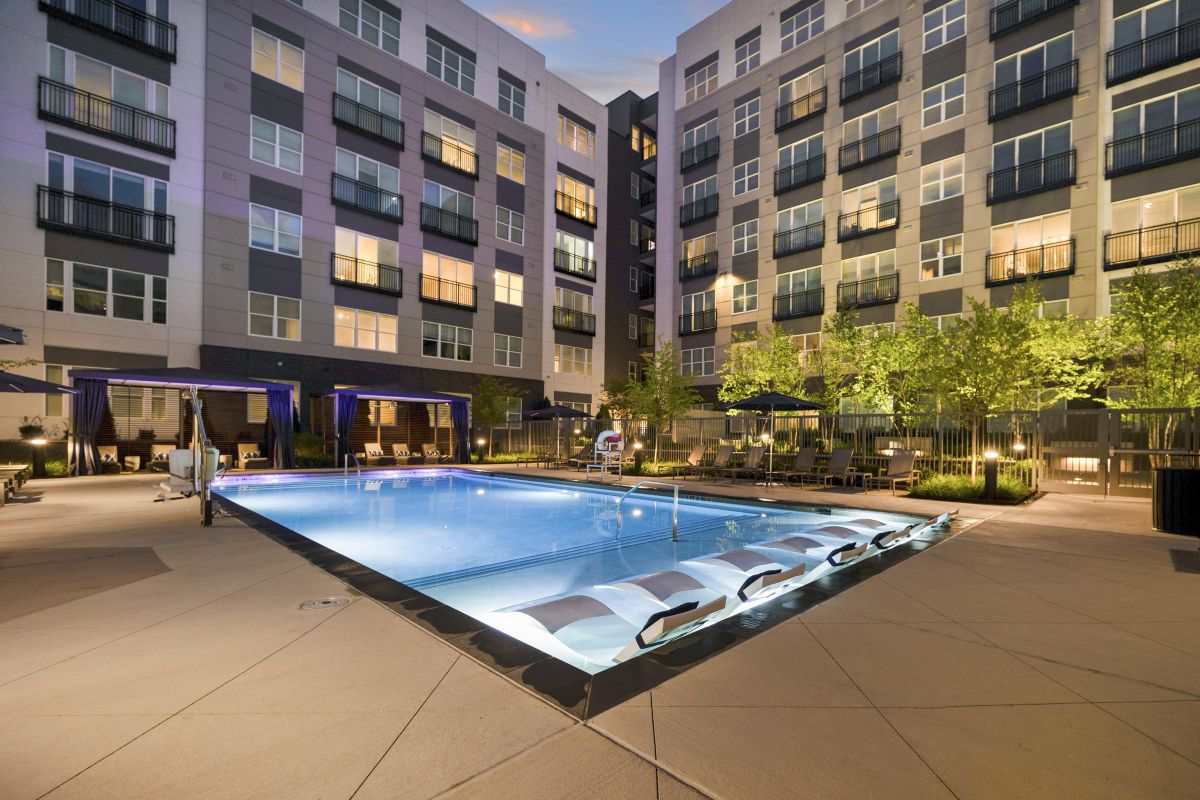 Modern apartment complex with a lit outdoor pool, surrounded by lounge chairs and cabanas at twilight.