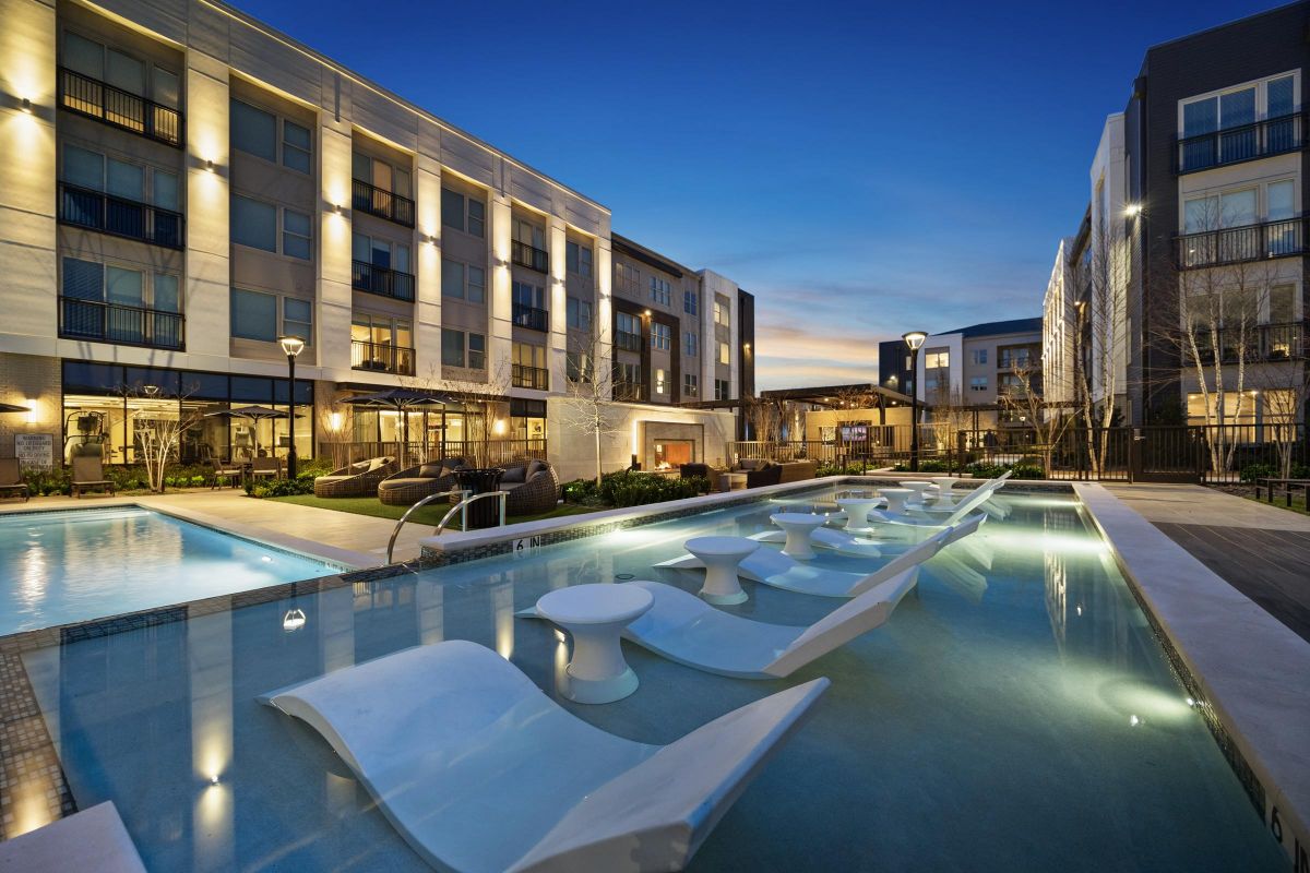 Modern apartment complex with a lit pool, lounge chairs in water, and seating areas at dusk.